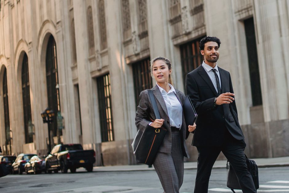 Two business professionals in suits walk down a city street beside a grand stone building, carrying briefcases and folders.