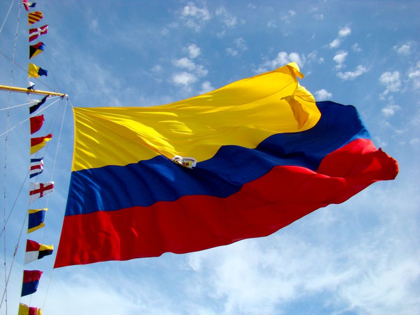 Colombian flag waving in the wind against a blue sky, showing yellow, blue, and red horizontal stripes with small flags on the left.