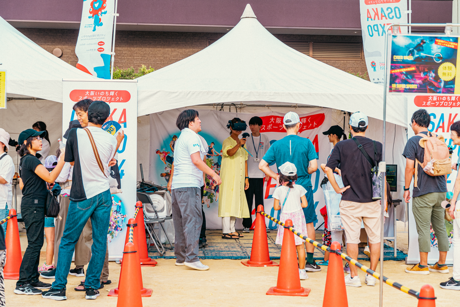 People line up at white tents for a daytime outdoor event, with orange traffic cones guiding the queue and banners in the background.
