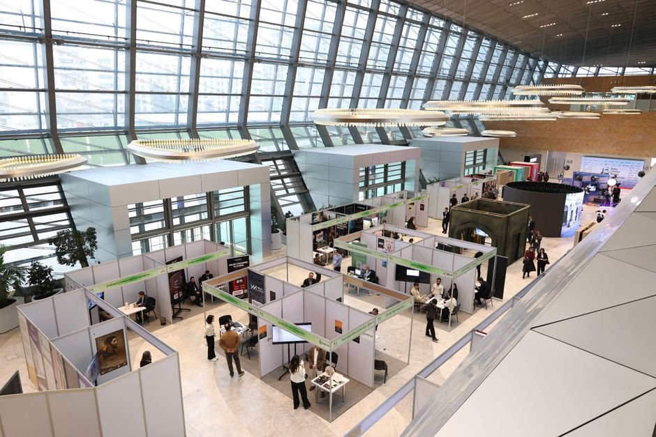 Exhibition hall with white booths and attendees at a trade fair inside a glass-walled atrium.