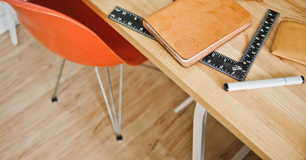 Desk setup with a tan leather notebook, black rulers, and a white marker on a light wood table; orange chair to the side.