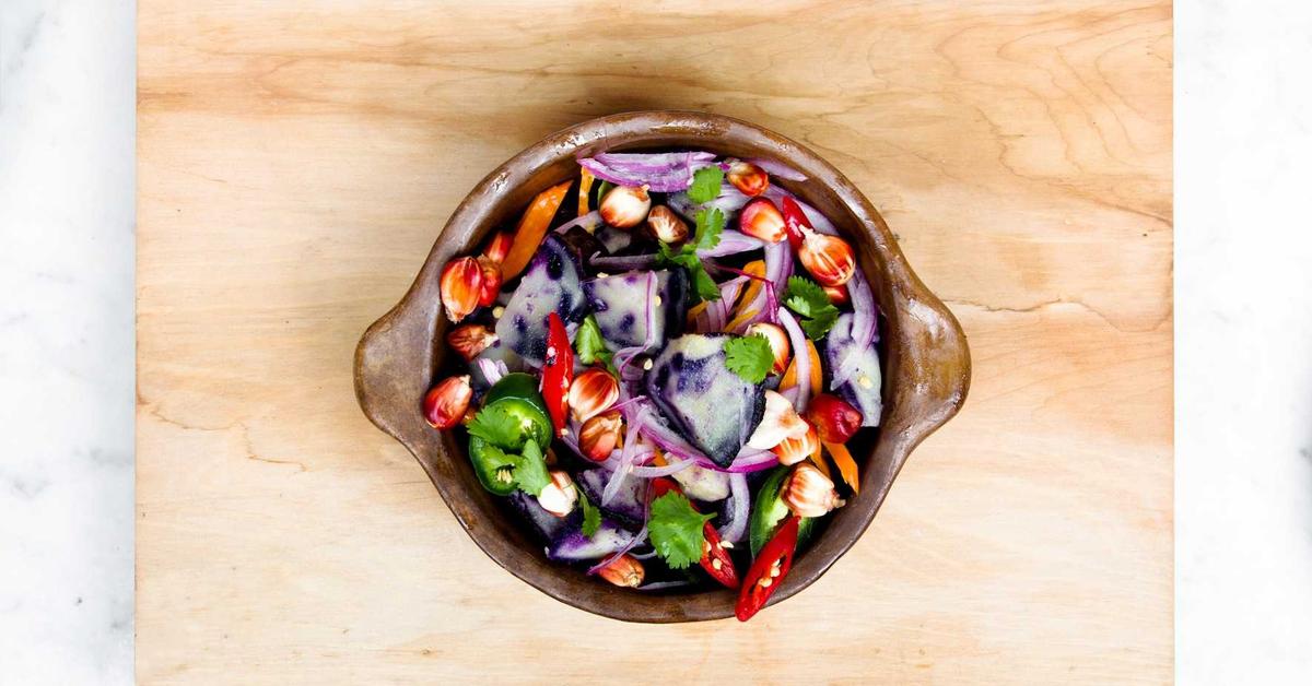 Colorful mixed salad in a brown ceramic bowl on a light wood board—purple potatoes, red onion, cilantro, and chilies.