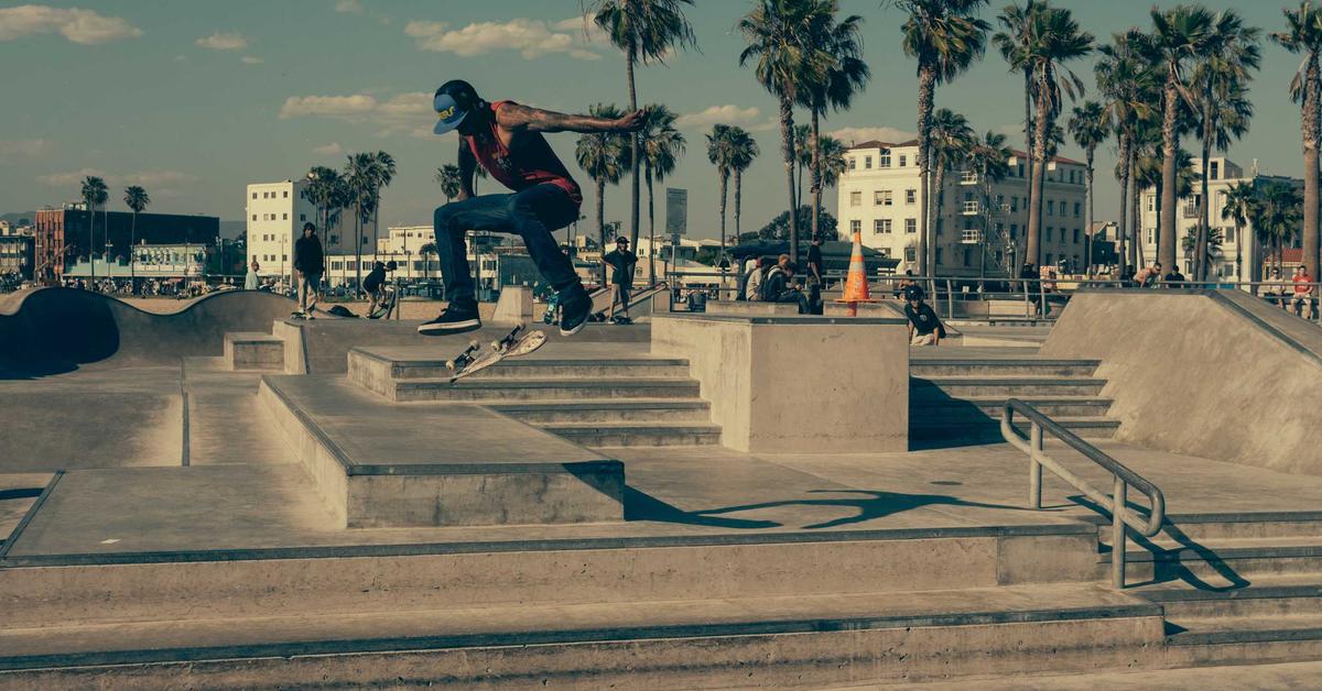 Skateboarder in mid-air performing a trick over a stair set at an outdoor skate park with palm trees in the background.