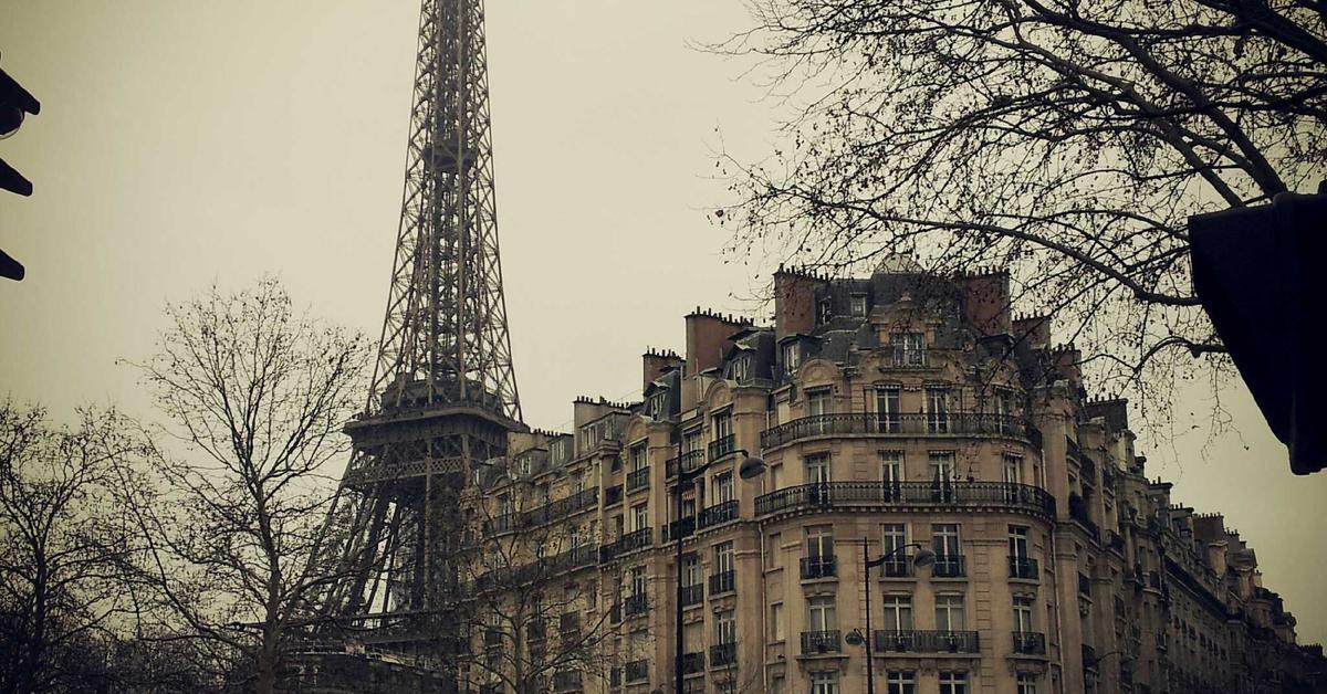Parisian Haussmann-style building with ornate balconies and the Eiffel Tower peeking behind on a cloudy day in Paris, France.