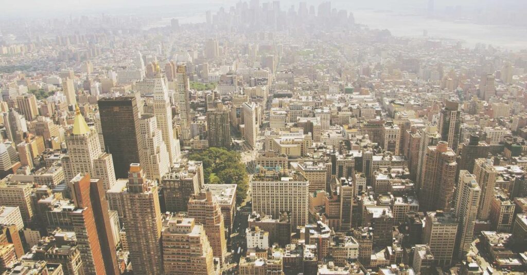 Aerial view of a dense urban cityscape with numerous high-rise buildings and grid-like streets in sunlight, viewed from above.