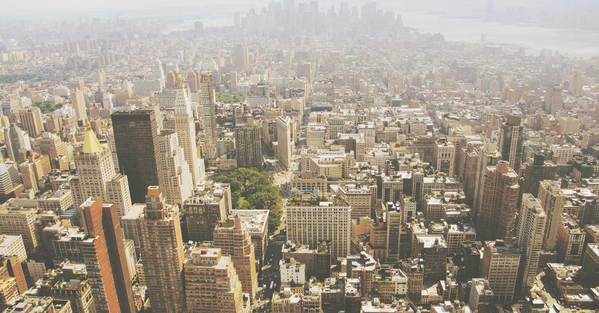 Aerial view of a dense urban cityscape with numerous high-rise buildings and grid-like streets in sunlight, viewed from above.