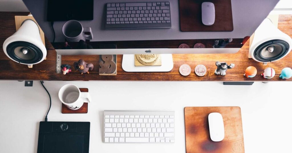 Top-down view of a modern desk setup: computer monitor, keyboard, mouse, white speakers, a mug, and small decorative figurines on a wooden shelf and desk imagery for a tidy workspace.