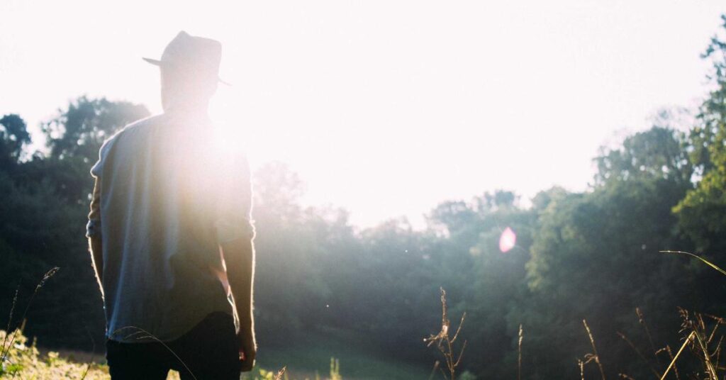 Person wearing a hat standing in a sunlit field, back to camera, early morning or late afternoon glow blocking part of the view.