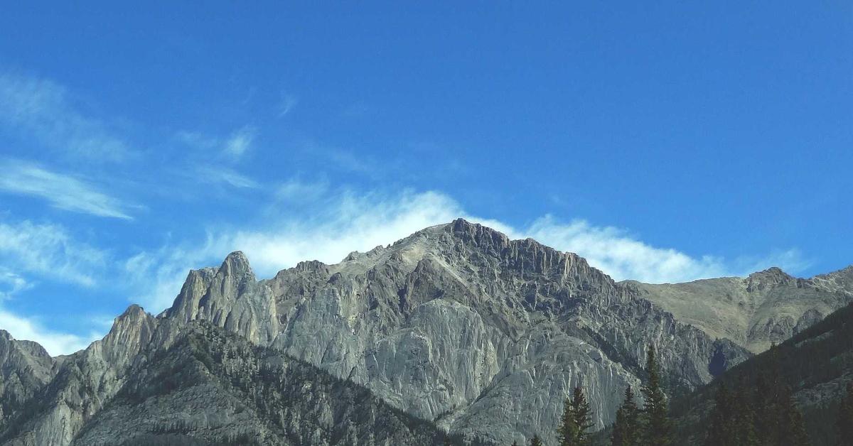 Rugged gray mountain peaks with a forested base beneath a clear blue sky and wispy clouds.