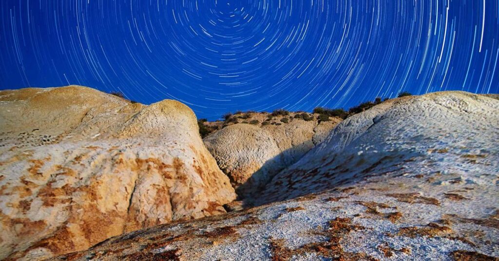 Night sky with circular star trails above rugged desert rocks and cliffs at dusk.