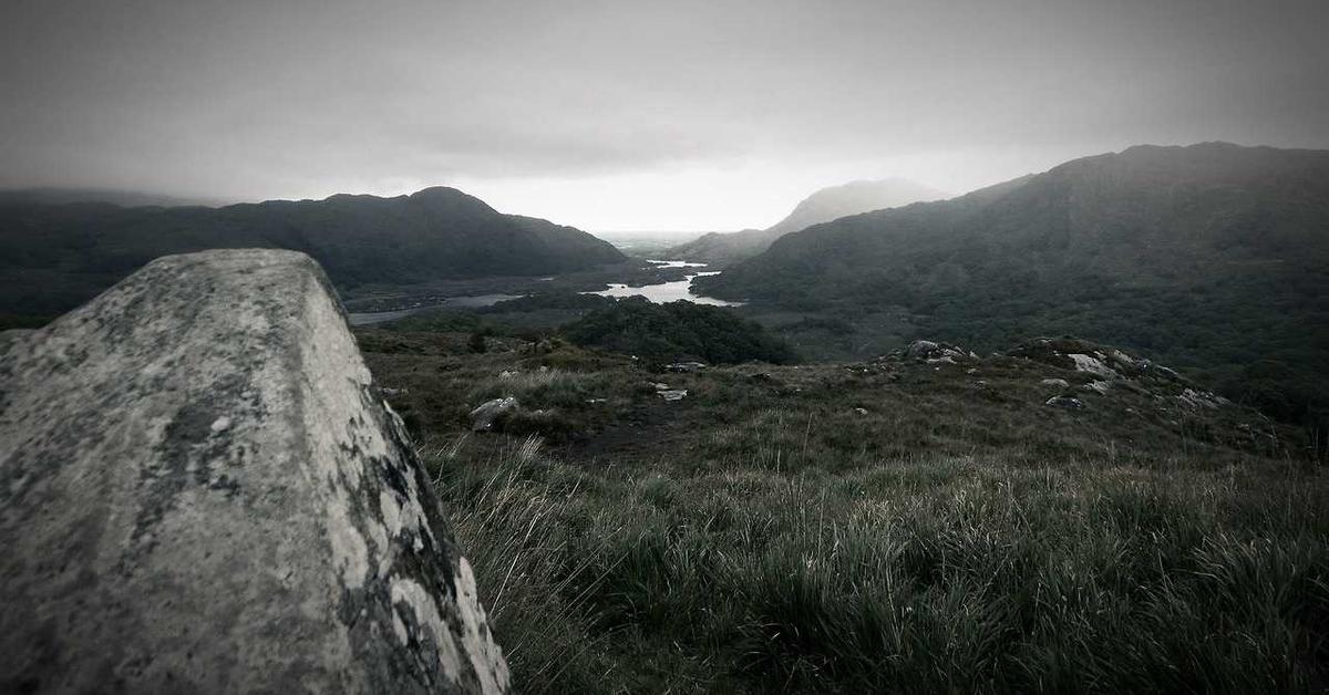 Moody valley landscape with a rocky foreground, rolling hills, and a winding river in the distance.