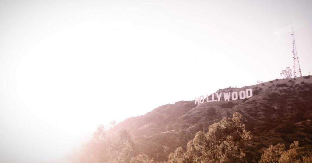 Hollywood sign perched on a sunlit hillside with trees in the foreground and a bright sky behind it.