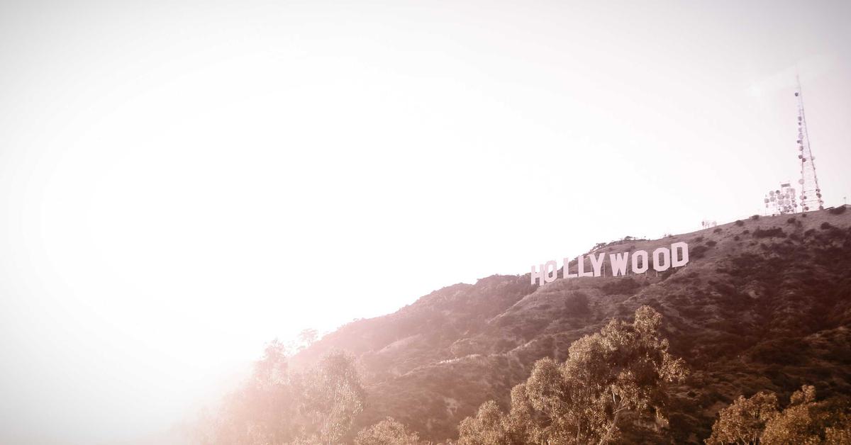 Hollywood sign perched on a sunlit hillside with trees in the foreground and a bright sky behind it.