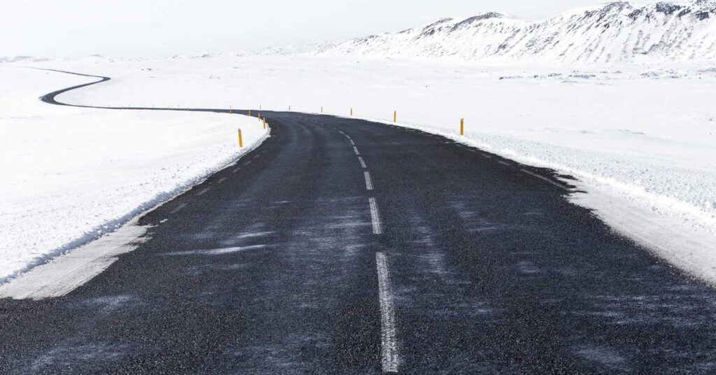 Snowy landscape with a winding asphalt road curving through white fields and distant hills, yellow roadside posts along the edge.
