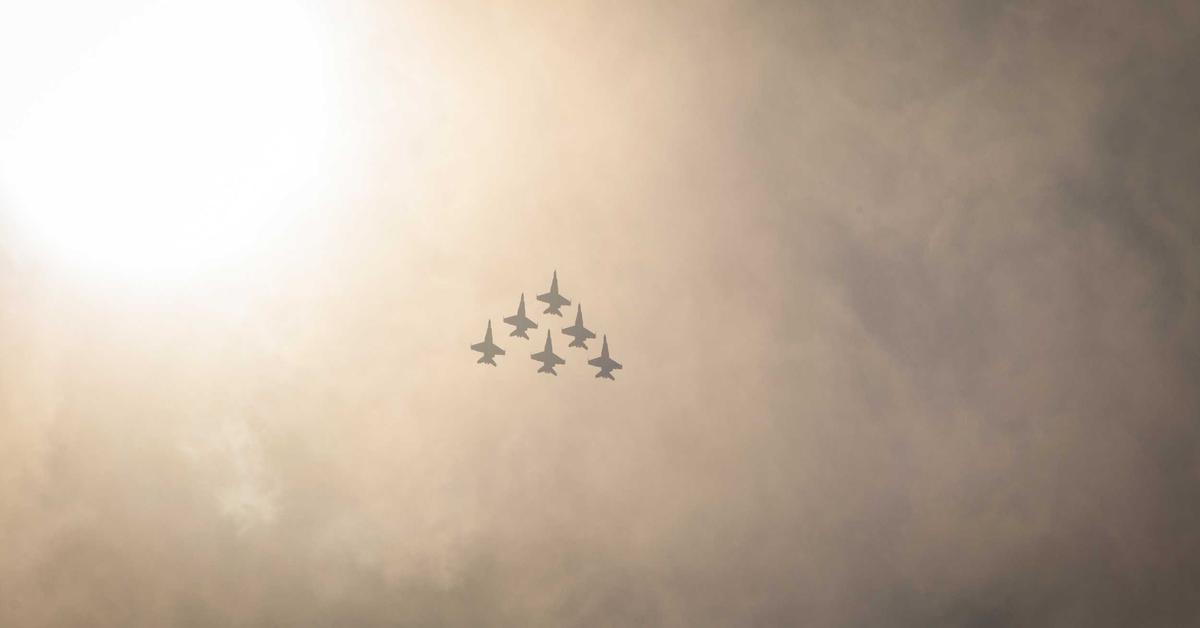 Six fighter jets in a tight formation flying across a hazy, sunlit sky.