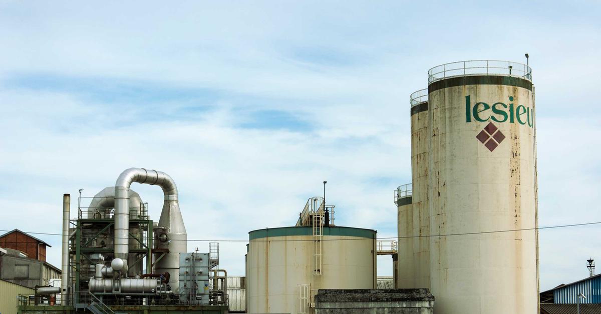 Industrial facility with a tall white silo marked 'lesieu' and an overhead network of silver pipes under a blue sky.