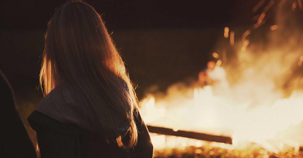Person with long hair seen from behind, standing near a large bonfire at night.