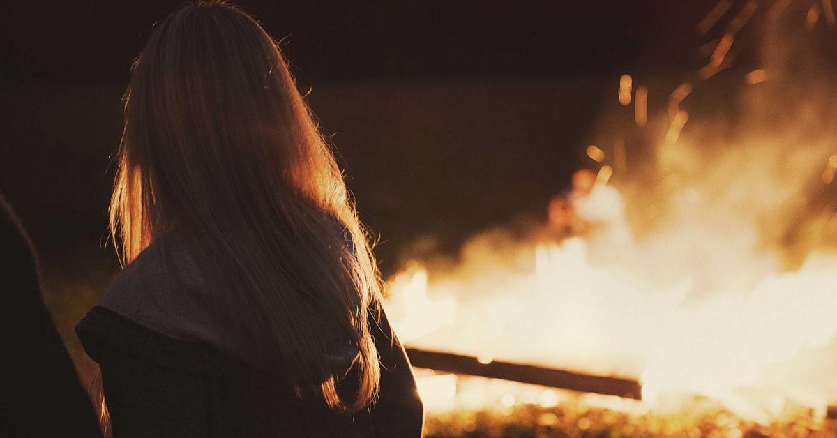 Person with long hair seen from behind, standing near a large bonfire at night.