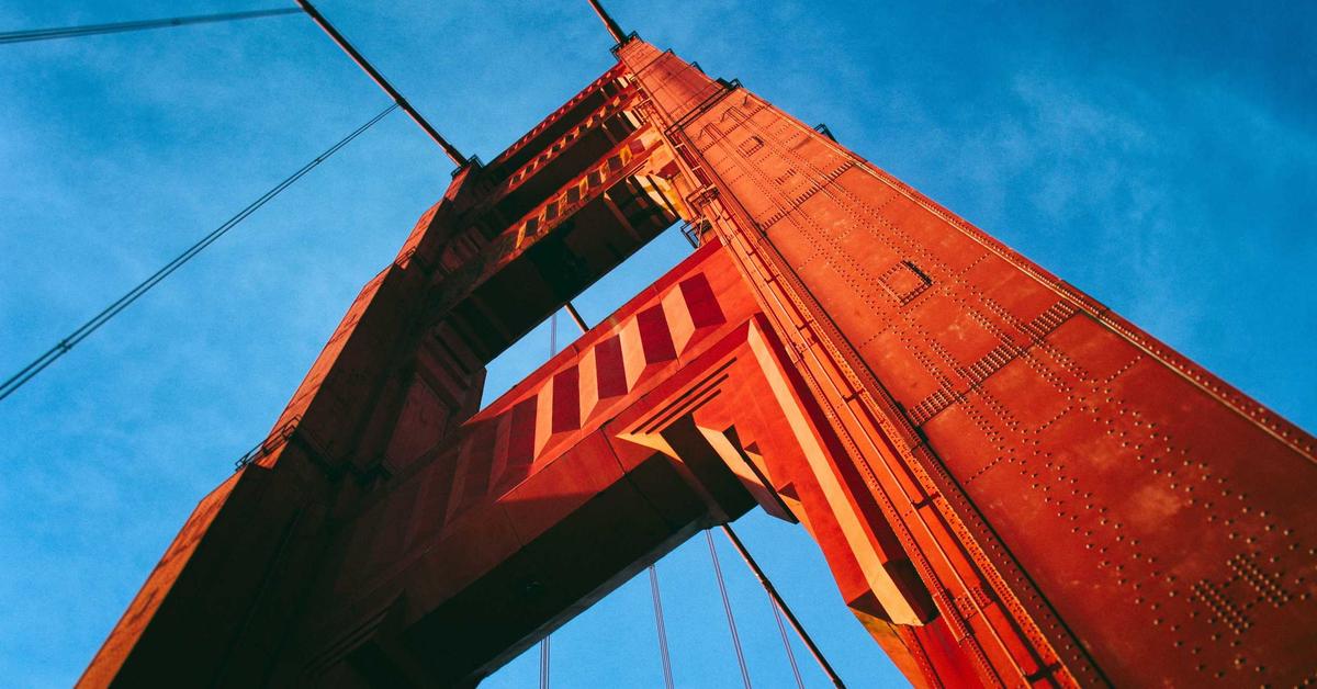 Close-up of a red suspension bridge tower with riveted steel beams against a bright blue sky, seen from below.