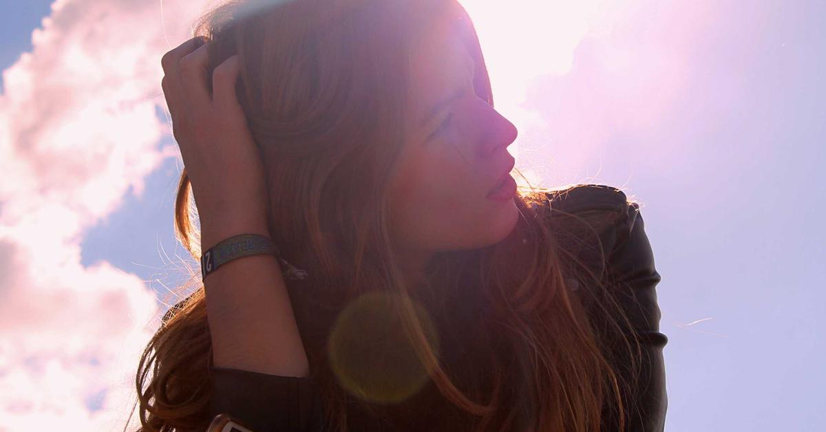Side-profile of a young woman outdoors, hand in hair, with bright sun and lens flare behind her.