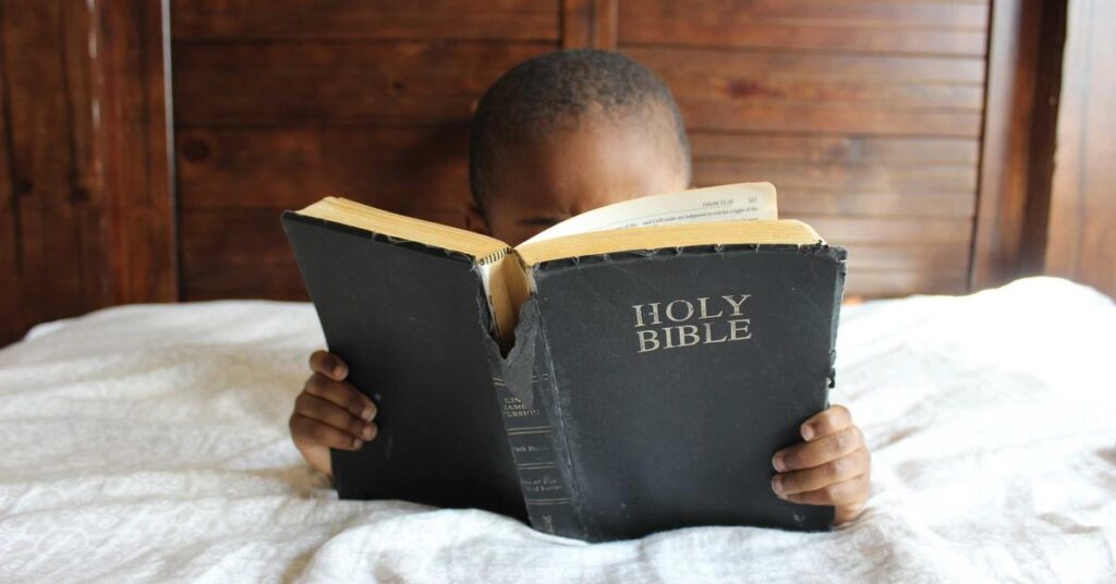 Child in bed reading a worn Holy Bible held open in front of their face.
