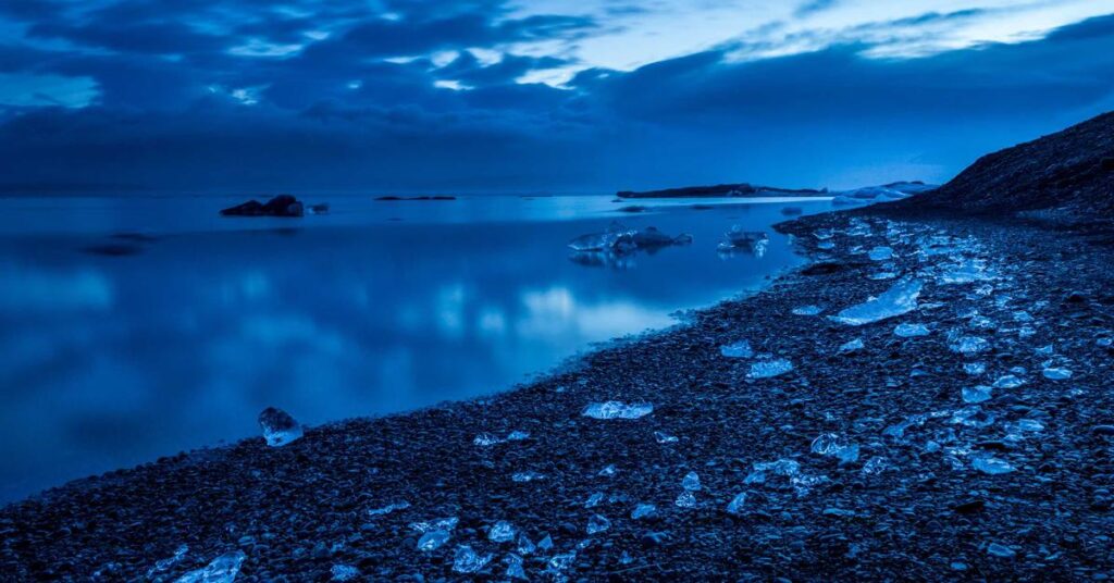 Blue-toned coastal scene with a rocky shoreline, scattered ice chunks, and calm water under a cloudy sky.