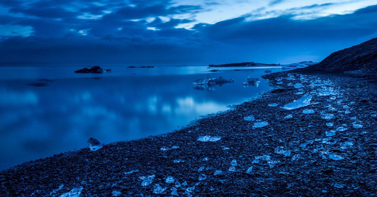 Blue-toned coastal scene with a rocky shoreline, scattered ice chunks, and calm water under a cloudy sky.
