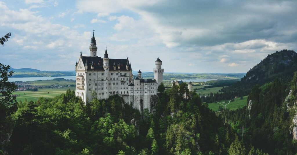 Neuschwanstein Castle perched on a forested hill with a valley and fields in the distance.