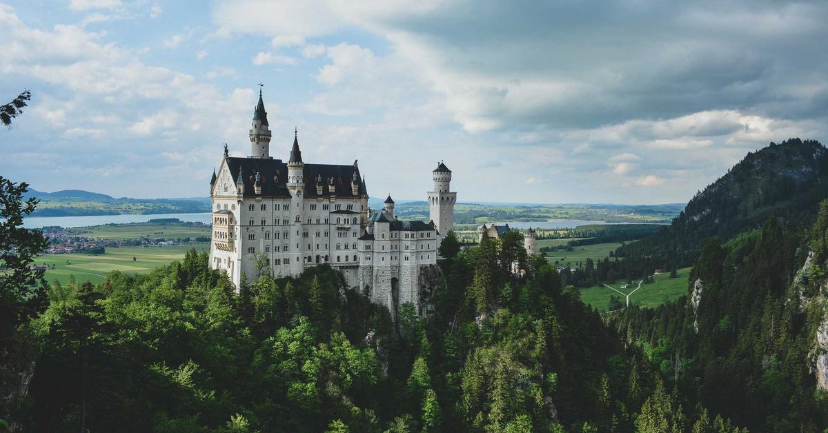 Neuschwanstein Castle perched on a forested hill with a valley and fields in the distance.