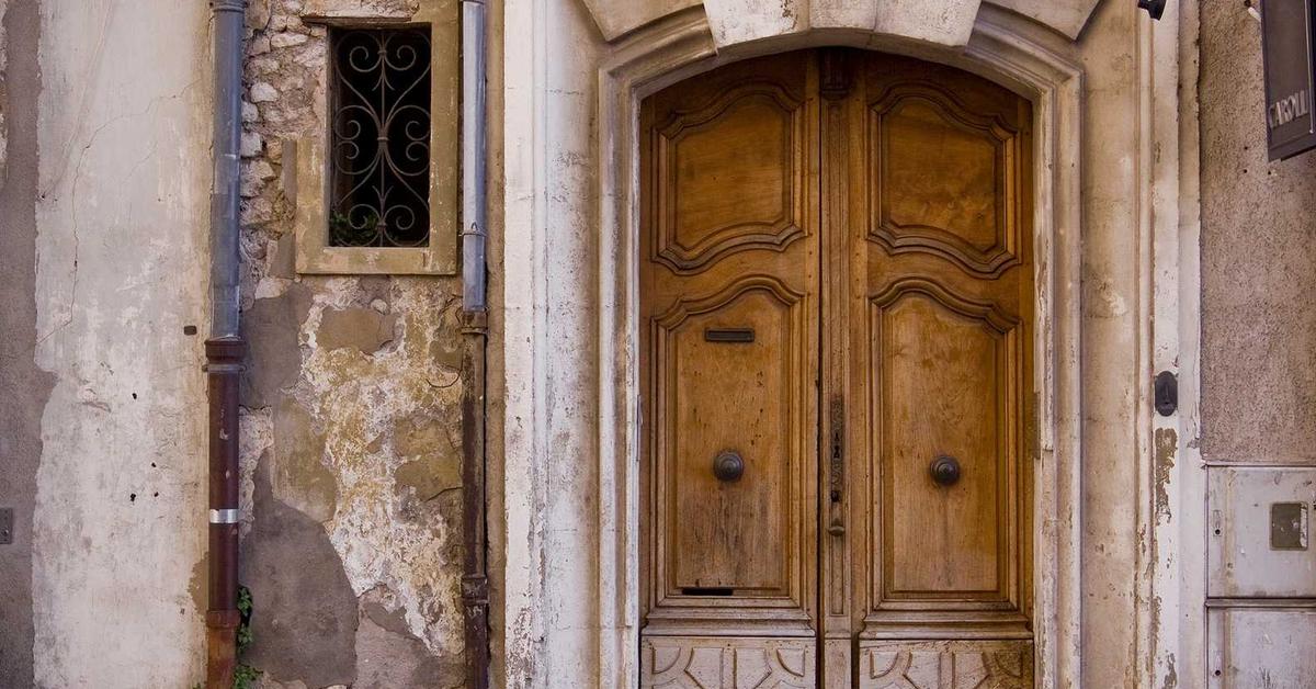 Weathered wooden double doors set in a stone building with peeling plaster and an arched stone frame by a narrow facade window with iron bars on the left.
