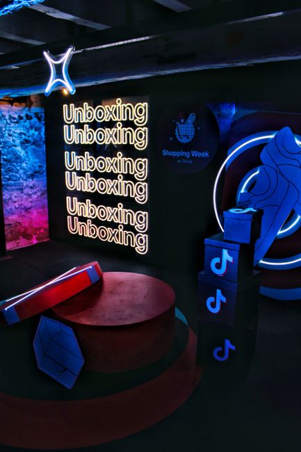 Dark exhibition booth with a neon 'Unboxing' sign repeated several times, blue neon rings, stacked TikTok logo blocks, and geometric props on a circular platform.