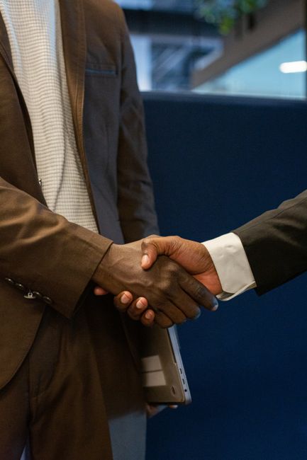 Two businesspeople shake hands in a professional setting; one in a brown suit holding a laptop, the other in a dark suit.