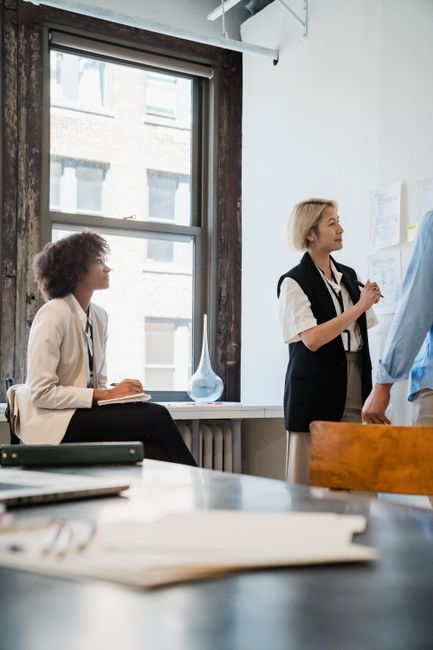Two women in a bright office; one stands at a whiteboard presenting, the other sits by a window taking notes.